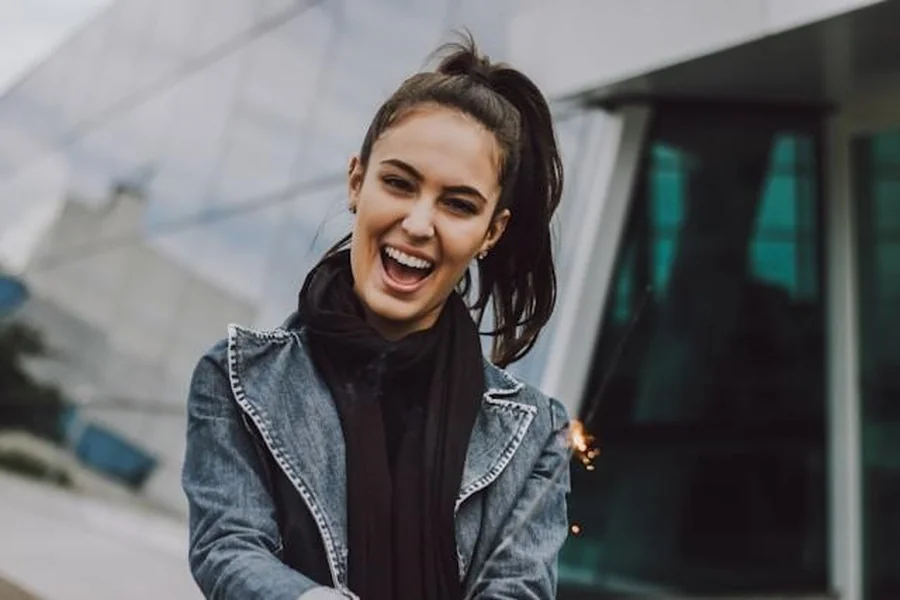 A young woman in a jean jacket and a black scarf, laughing in front of a modern building.